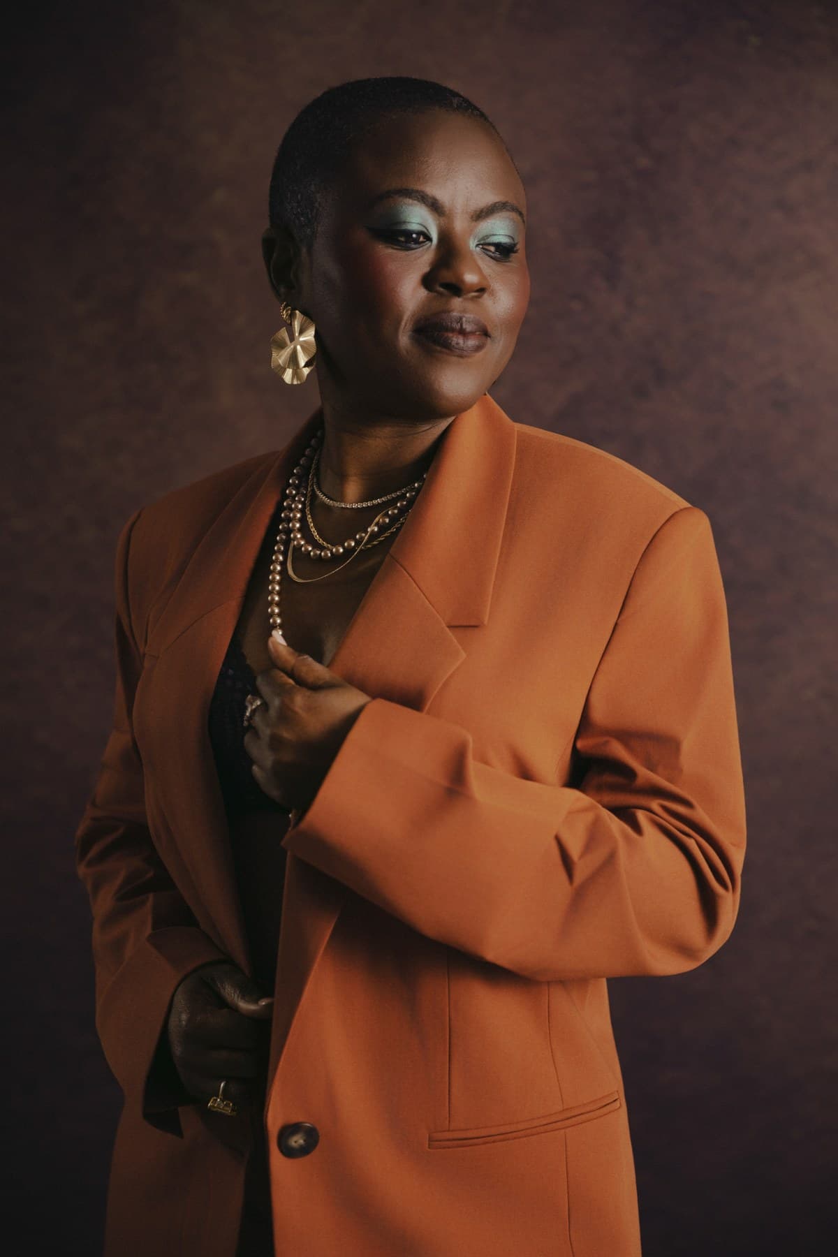 A woman in an orange blazer with bold eyeshadow and gold earrings, a striking studio portrait at Body & Soul Studios.