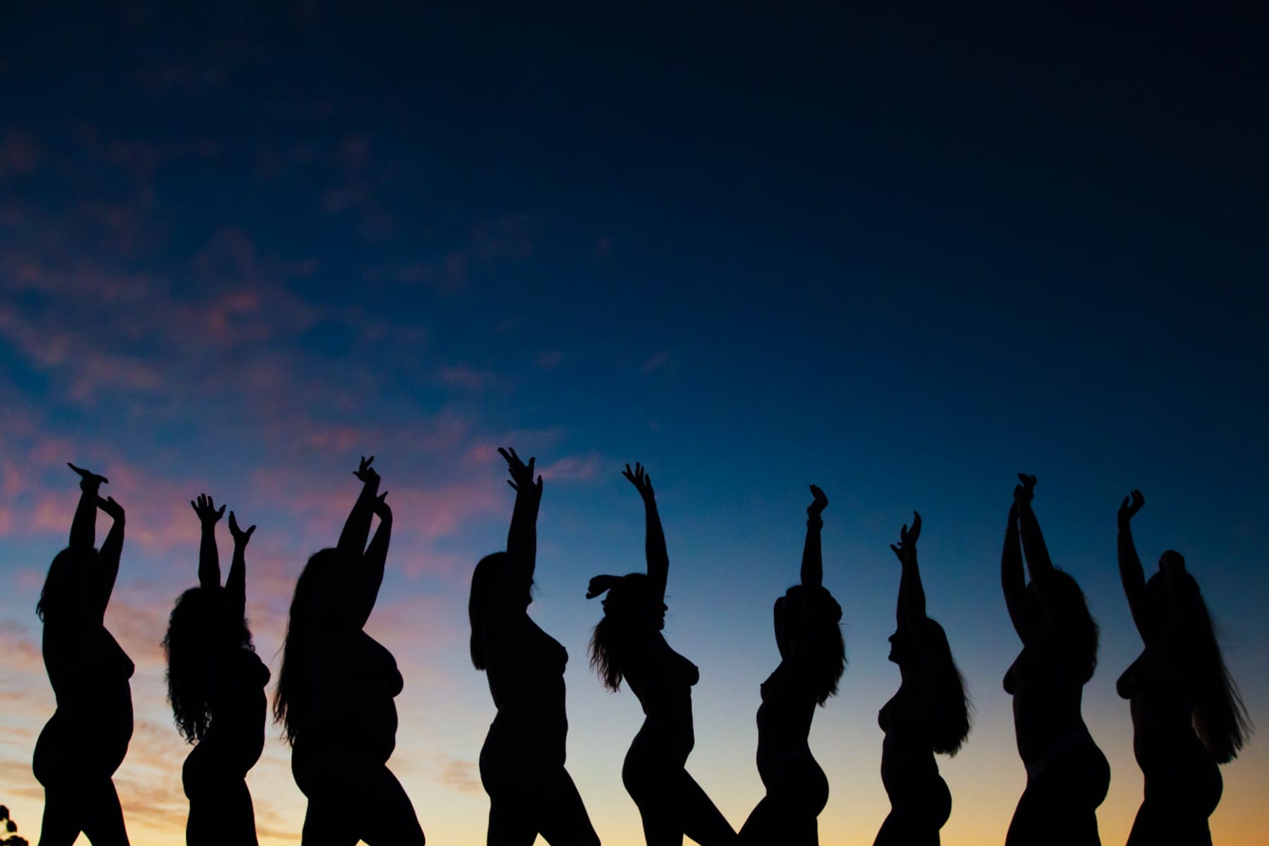 Silhouettes of women with arms raised against a sunset sky, a Body & Soul gathering.