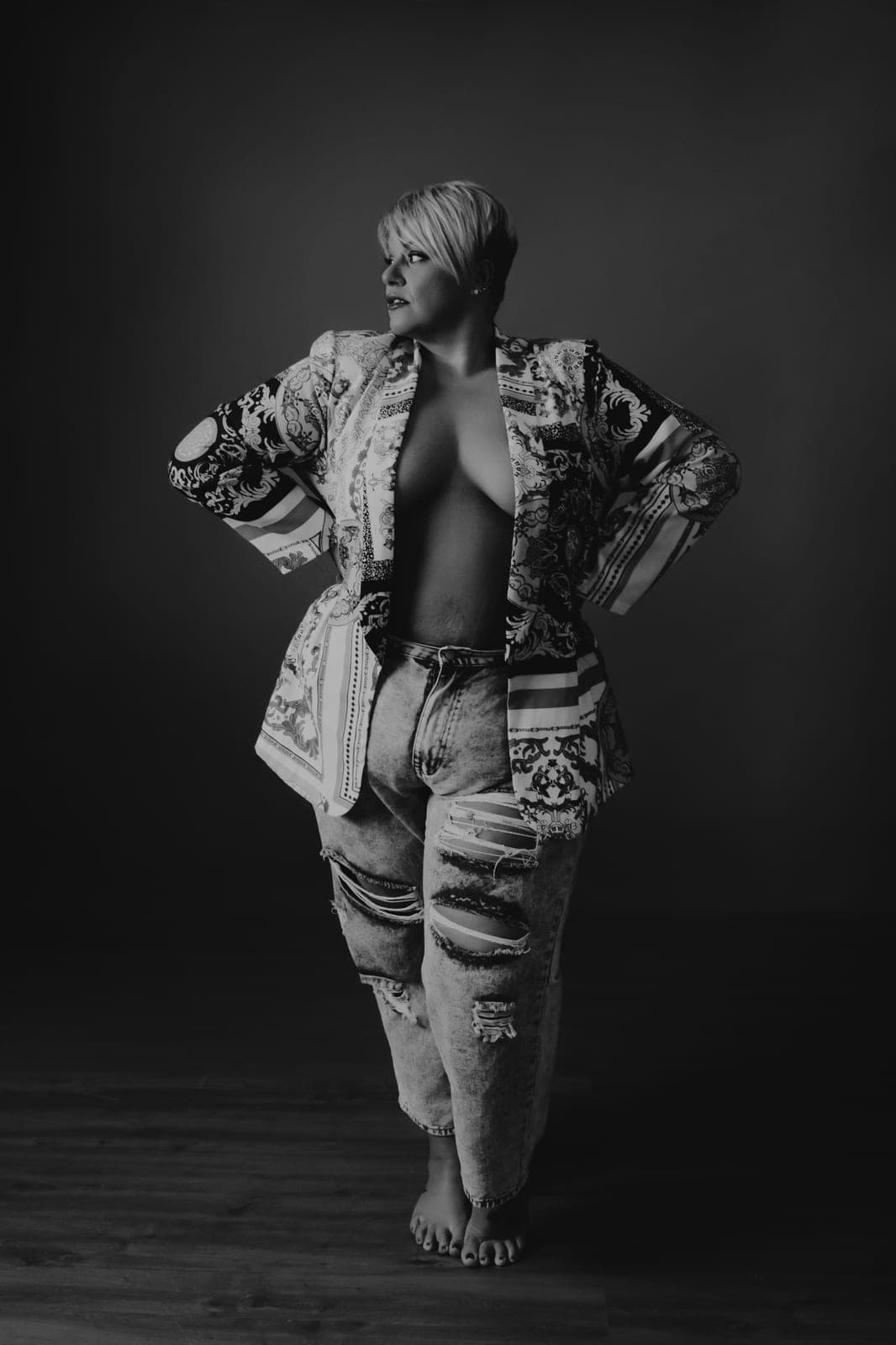Curvy woman in a baroque-print blazer and ripped jeans, standing tall and barefoot, a striking black-and-white portrait at Body & Soul Studios, London.